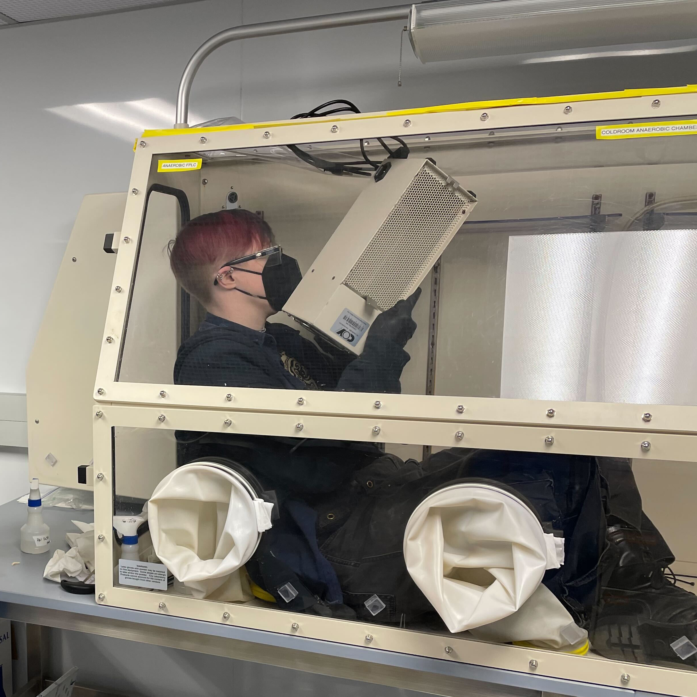 a photo of dr. g. e. kenney sitting inside a glovebox in a cold room and lifting a piece of equipment into place.